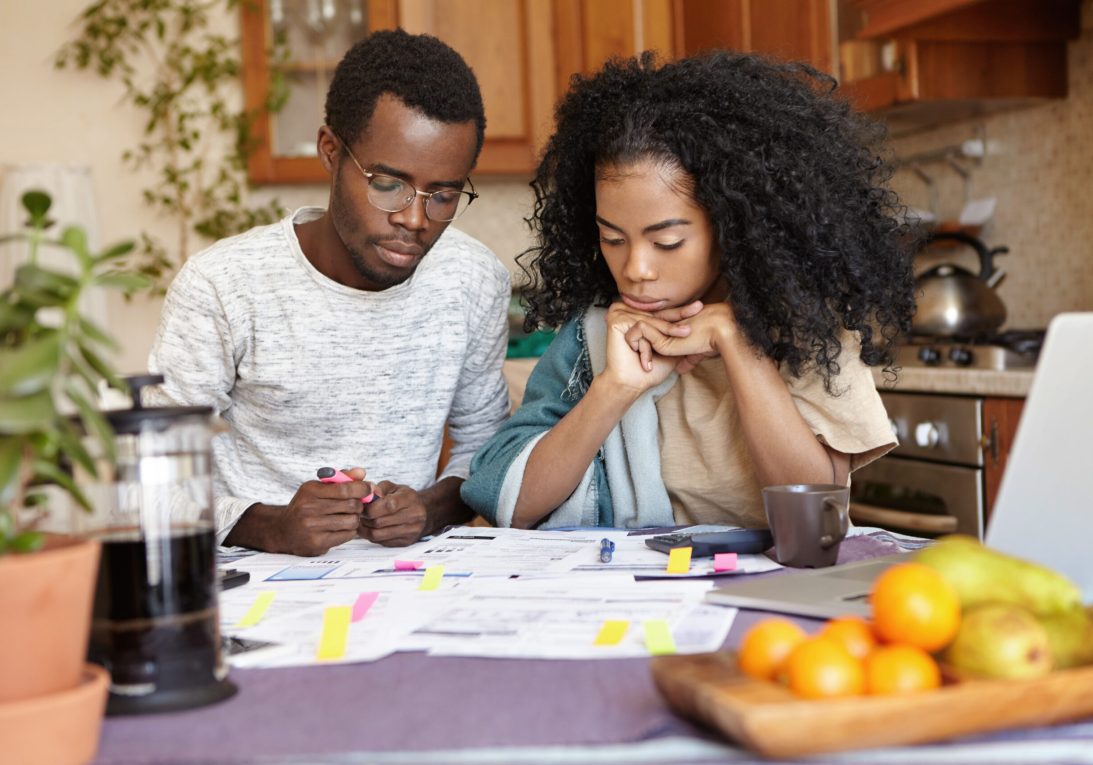 Young African couple doing paperwork together, sitting at kitchen table with lots of papers, calculator and laptop, looking frustrated. Dark-skinned family calculating domestic budget at home